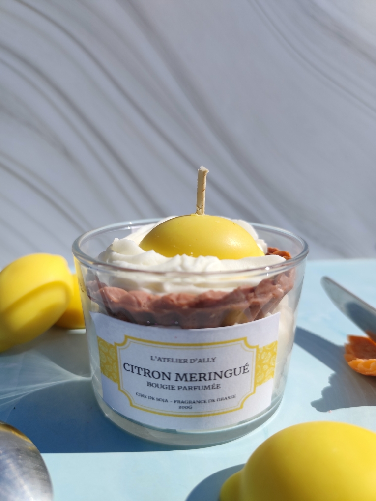 Bougie Citron Meringuée dans pot en verre avec gâteau en cire sur chantilly blanche, coulis jaune citron, crème pâtissière, citron entier au centre, citrons en cire et cuillère à côté.