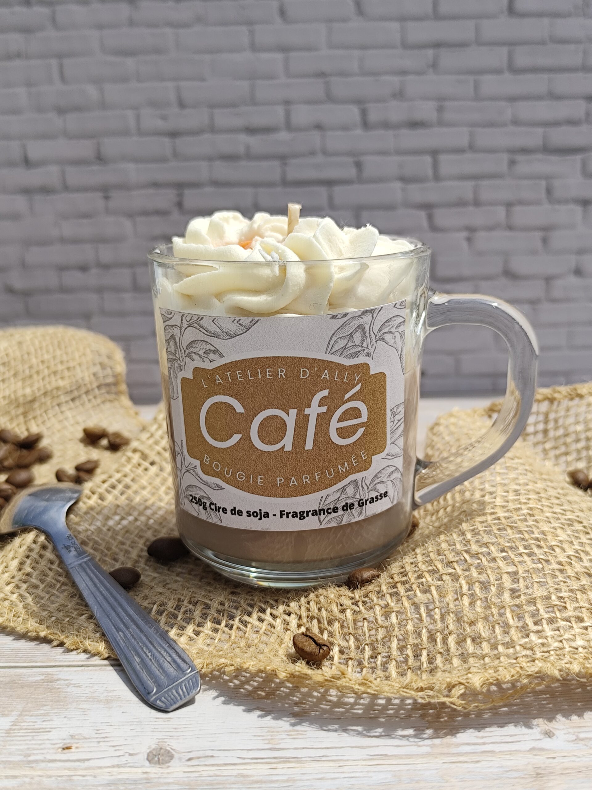 Bougie Café dans un pot en verre avec base marron, chantilly blanche et grains de café en cire. Cuillère à café posée à côté et grains de café dispersés autour.