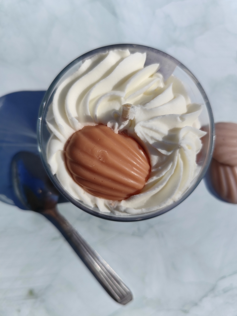 Vue de dessus de la bougie Madeleine avec chantilly blanche, madeleine en cire au centre, cuillère à côté et madeleines en cire disposées autour.