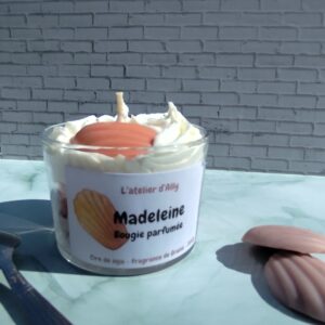 Bougie Madeleine dans pot en verre, chantilly blanche et madeleine en cire posée au sommet. Cuillère placée à côté de la bougie.