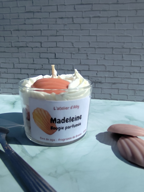 Bougie Madeleine dans pot en verre, chantilly blanche et madeleine en cire posée au sommet. Cuillère placée à côté de la bougie.