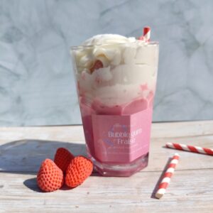 Bougie Bubblegum Fraise dans un pot en verre, base rose, chantilly blanche avec fraises en cire, paille rouge et blanche et paillettes argentées. Décor avec pailles et fraises en cire autour.