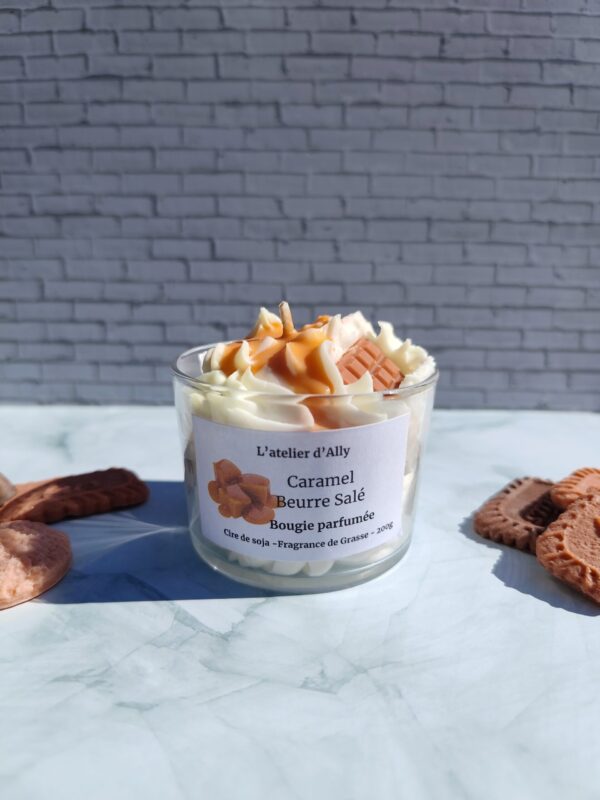 Bougie Caramel Beurre Salé dans pot en verre, avec chantilly blanche, coulis de caramel et tablette en cire couleur caramel. Décor gourmand avec biscuits autour.
