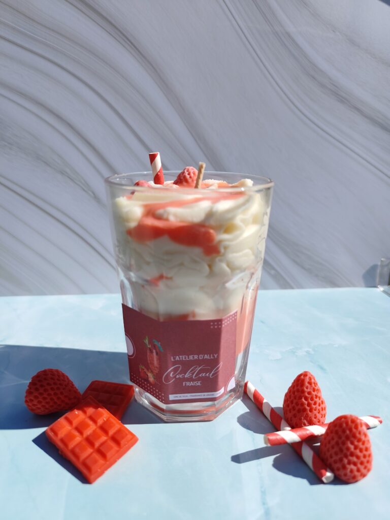 Bougie Cocktail Fraise dans son grand pot en verre, chantilly blanche, coulis rouge et fraise en cire avec paille rouge et blanche. Décor avec fraises en cire et tablette de chocolat rouge.