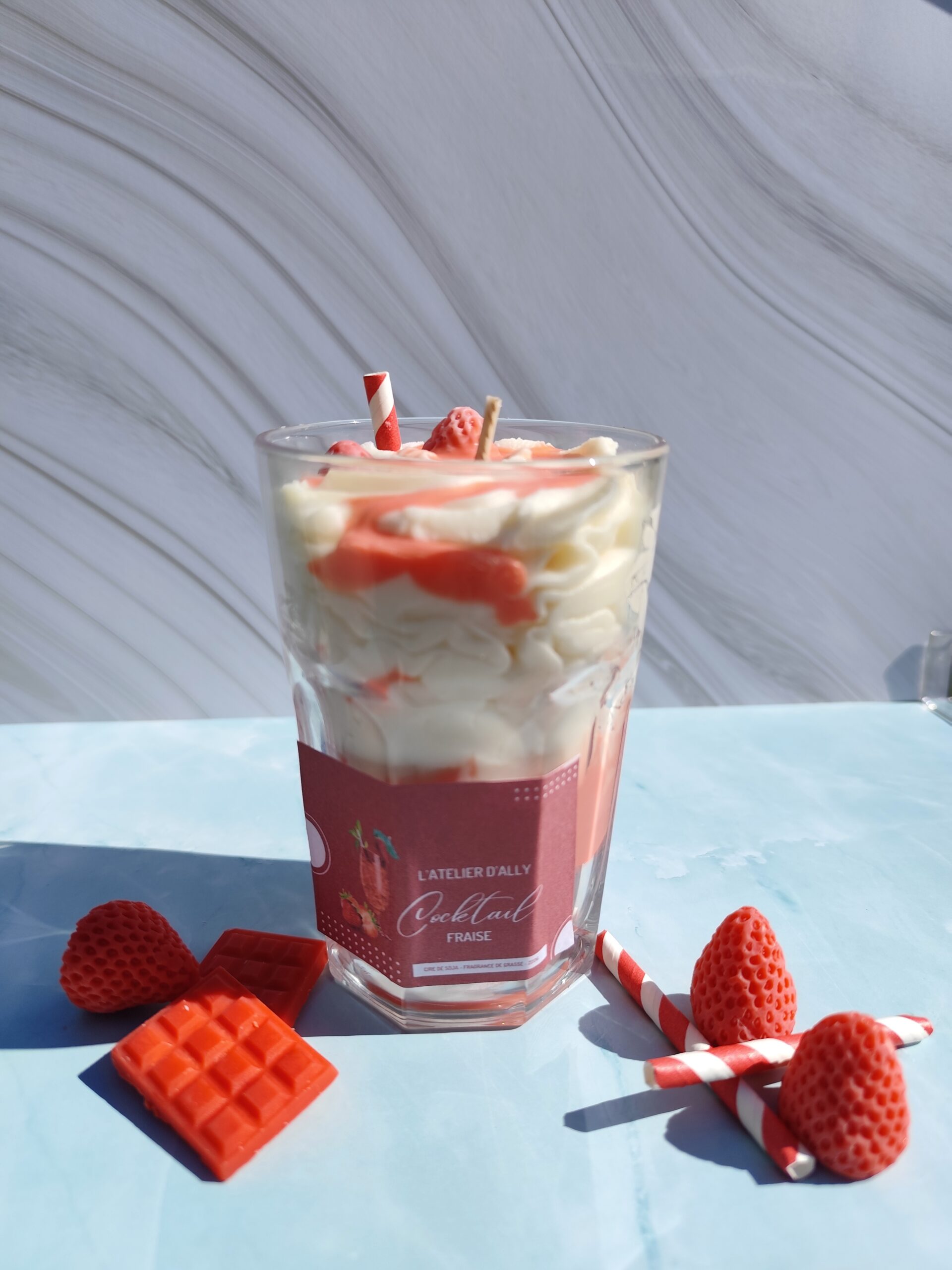 Bougie Cocktail Fraise dans son grand pot en verre, chantilly blanche, coulis rouge et fraise en cire avec paille rouge et blanche. Décor avec fraises en cire et tablette de chocolat rouge.
