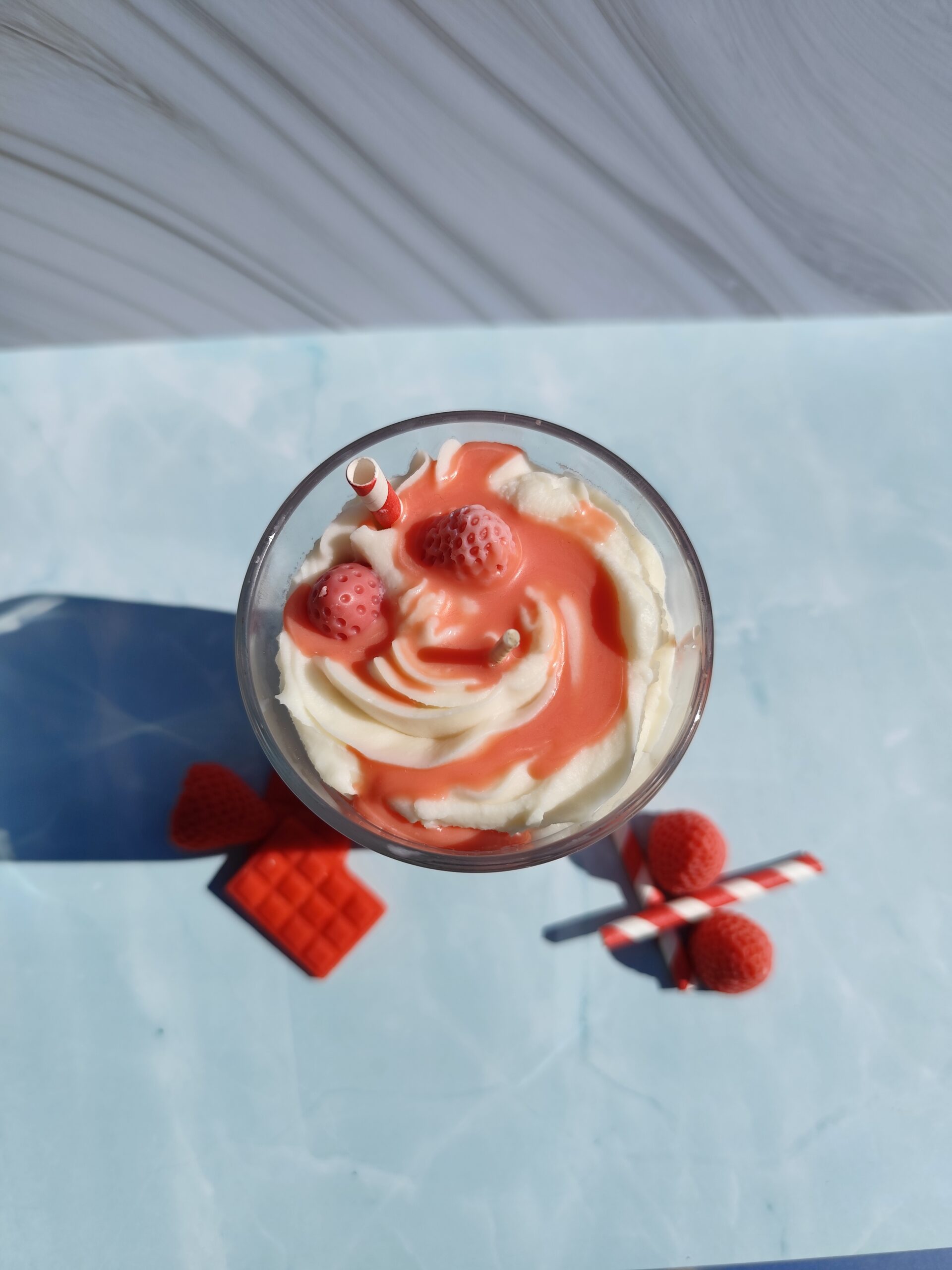 Vue de dessus de la bougie Cocktail Fraise avec chantilly blanche, coulis rouge, fraise en cire et paille rouge est blanche. Décor avec fraises en cire et tablette de chocolat rouge.