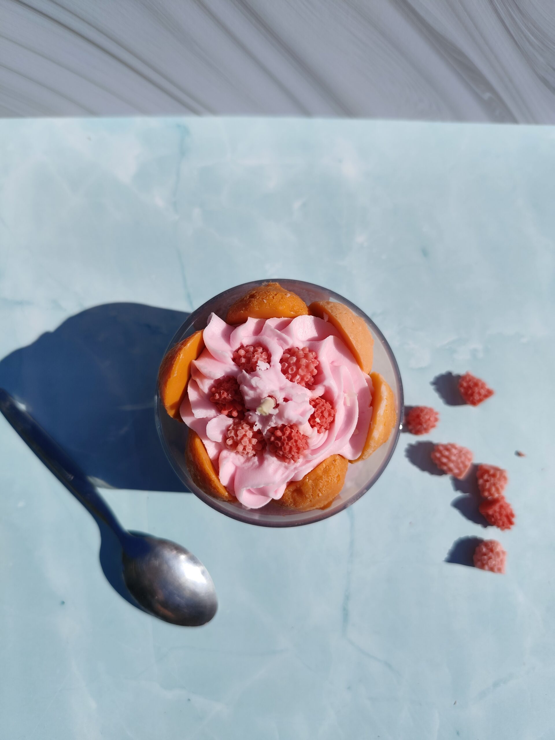 Vue de dessus de la bougie Charlotte aux framboises avec framboise en cire au centre et biscuits à la cuillère en cire autour. Framboises décoratives et cuillère à côté.