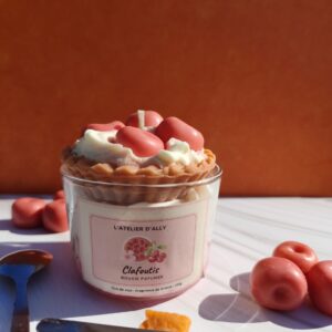 Bougie Clafoutis sur fond orange foncé, avec décor ultra réaliste : gâteau en cire, chantilly blanche, coulis rouge et cerises, mise ne scène avec couteau, biscuit et cerises autour