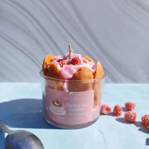 Bougie Charlotte aux framboises dans pot en verre, entourée de biscuits à la cuillère en cire, chantilly rosée et framboises en cire au centre. Décor avec framboises en cire et cuillère.