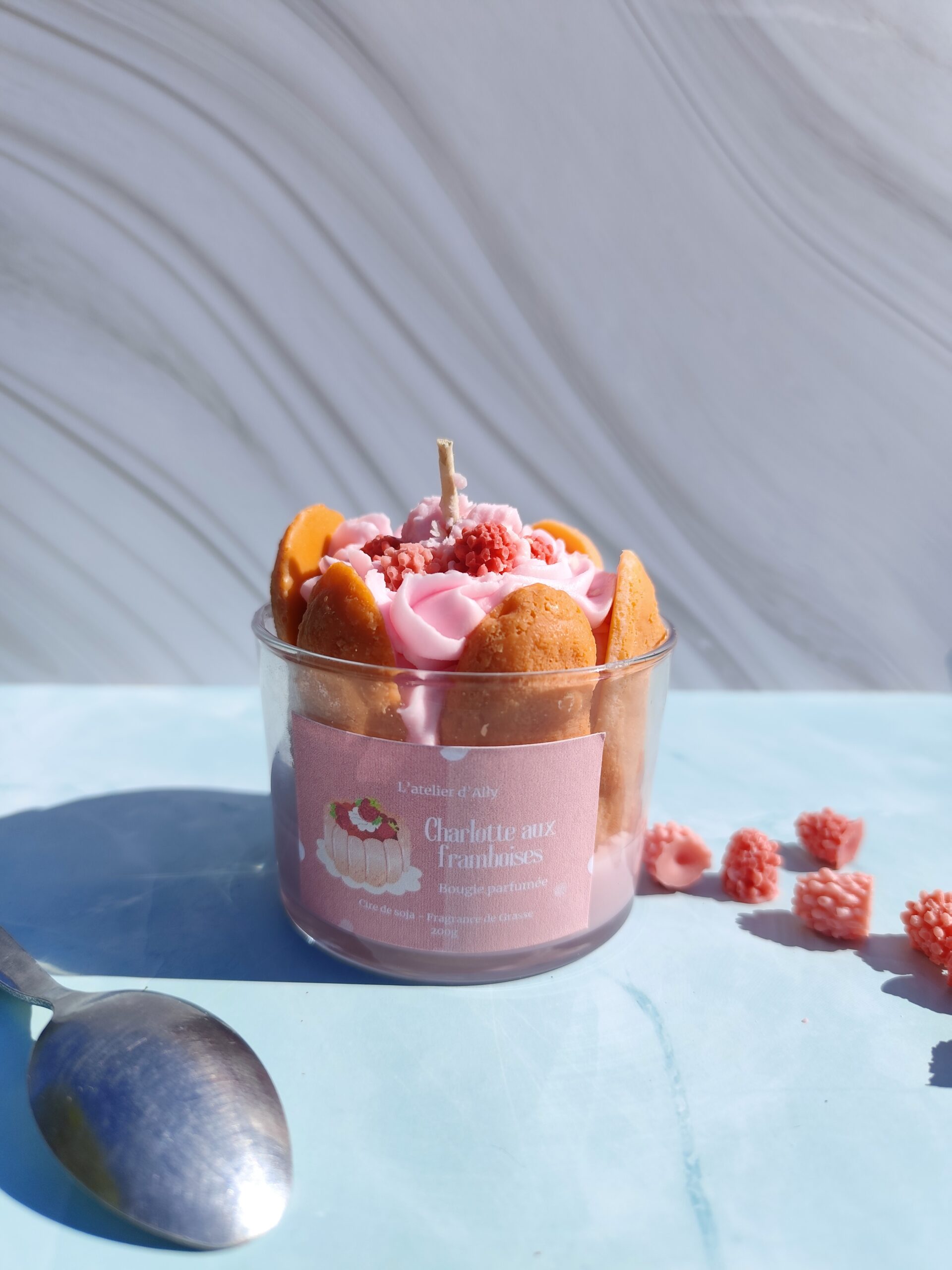 Bougie Charlotte aux framboises dans pot en verre, entourée de biscuits à la cuillère en cire, chantilly rosée et framboises en cire au centre. Décor avec framboises en cire et cuillère.