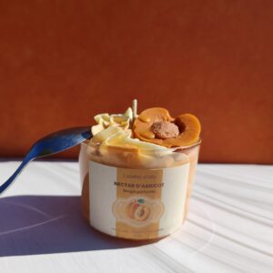 Bougie gourmande Nectar d'abricot avec chantilly blanche, coulis orange et demi- abricot en cire, posée devant un fond orange avec cuillère sur le pot