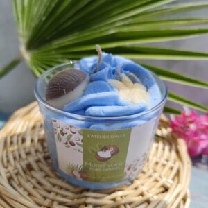 Bougie Monoï Coco sur panier en osier avec feuille de palmier, chantilly bleue, décor noix de coco dorée et fleur de tiaré blanche dorée, étiquette exotique visible.