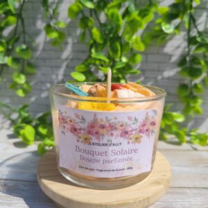Bougie Bouquet Solaire 200g dans son pot en verre recyclable, vue de face, chantilly orange et fleurs en cire avec paillettes dorées, décor estival en fond
