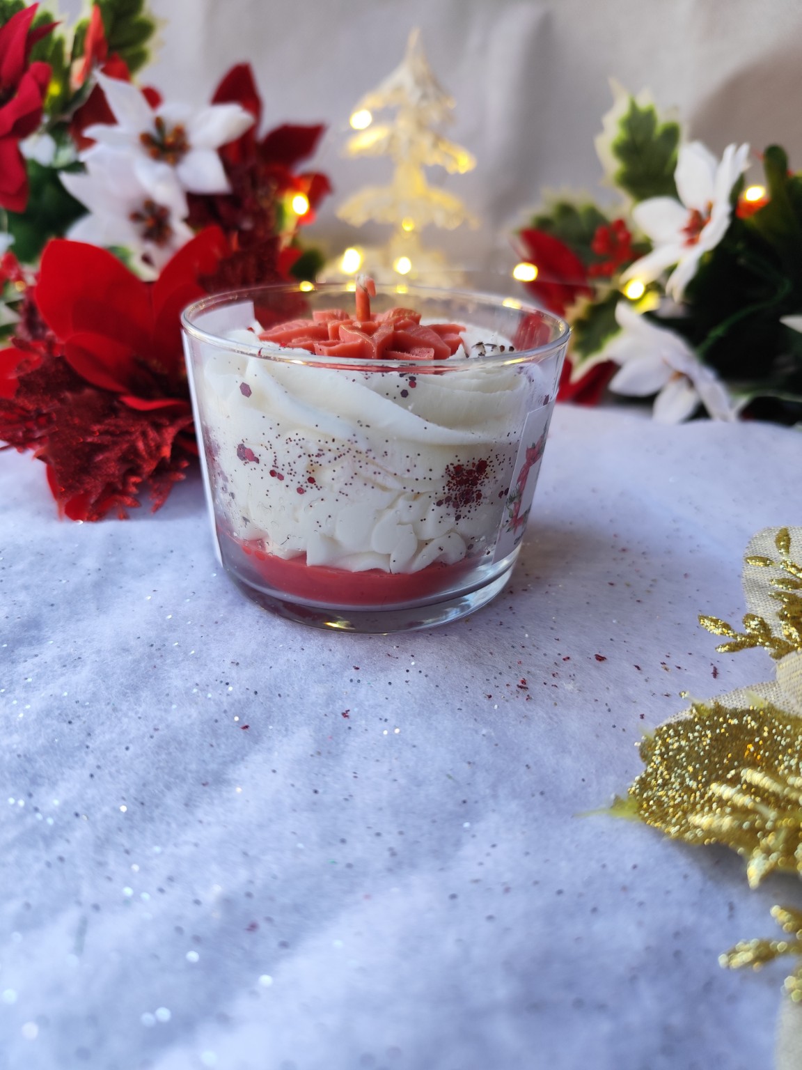 Bougie artisanale Fleur de Noël dans un décor de fête avec fleurs rouges et blanches et fond pailleté.