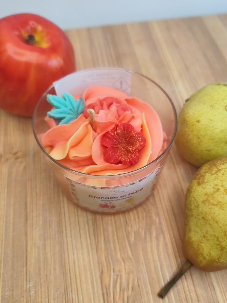 Vue de dessus de la bougie artisanale Grenade et Poire avec sa chantilly bicolore rouge et jaune, sa fleur rouge et sa feuille verte, entourée d'une pomme et de poires.