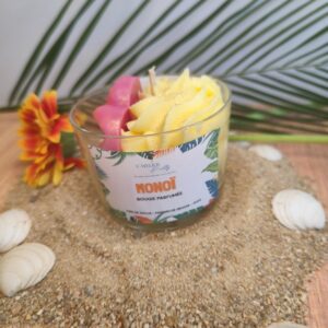 Bougie Monoï artisanale décorée d'une fleur exotique jaune et rose, mise en scène sur un décor de plage avec sable, galets et feuille tropicale.